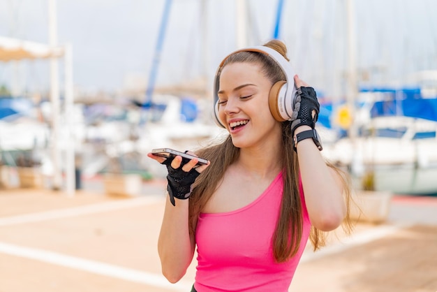 Chica joven bonita del deporte al aire libre escuchando música con un móvil y cantando