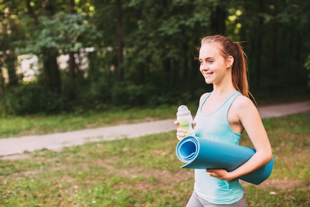 Chica joven con una botella de agua y colchoneta de gimnasia en el parque