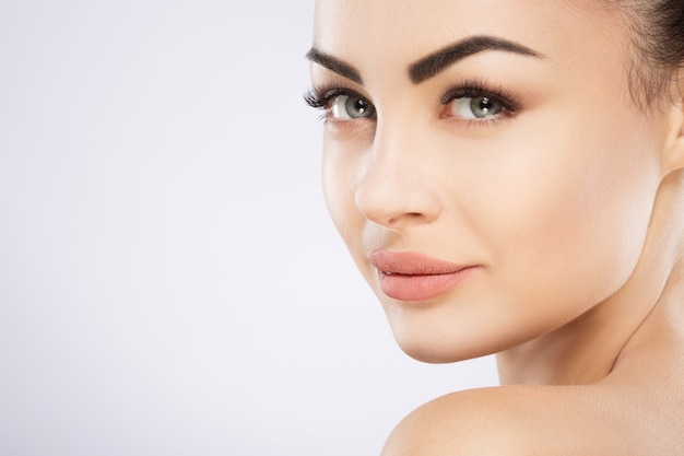 Chica joven con cabello negro fijo detrás, ojos grandes, cejas gruesas y hombros desnudos en la pared gris, de cerca.