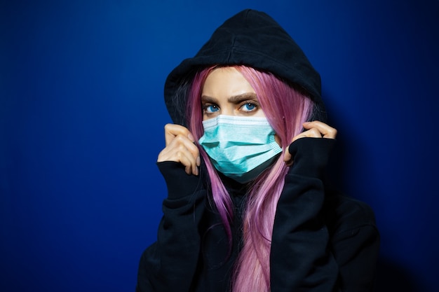 Chica joven con cabello rosado y ojos azules, con máscara médica contra la gripe en un suéter con capucha en la pared de color azul oscuro.