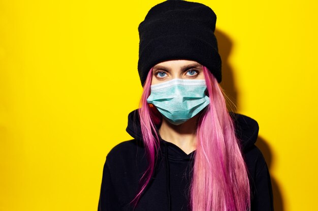 Chica joven con cabello rosado y ojos azules, con máscara médica contra la gripe, vestida con un suéter con capucha negro y un gorro en la pared amarilla.