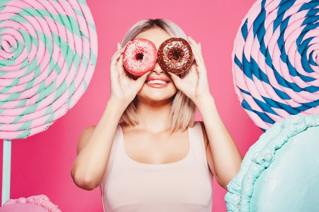 Chica joven con cabello rubio vistiendo top y gorra rosa de pie con enormes paletas dulces en pink studio