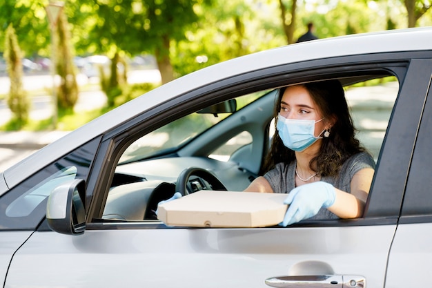 Chica joven con caja de pizza en coche con máscara médica y guantes