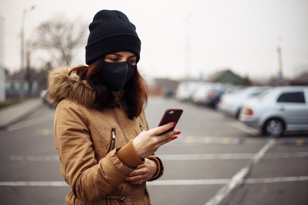 Chica joven en las calles de la ciudad vistiendo una mascarilla médica estéril negra. Mujer que usa el teléfono para buscar noticias sobre nCov 2019. Concepto de atención médica y epidemia de coronavirus pandémico COVID-19 en cuarentena.