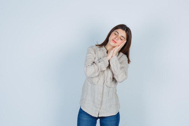 Chica joven en camisa beige, jeans apoyado en la mejilla en las palmas de las manos como almohada y mirando soñoliento, vista frontal.