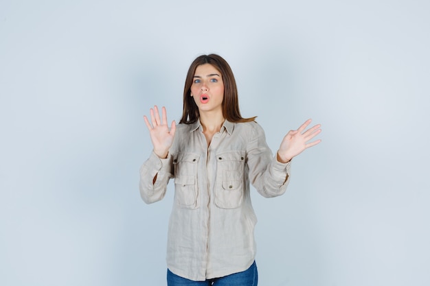 Chica joven con camisa beige, jeans levantando las manos como mostrando un gesto de restricción y mirando sorprendido, vista frontal.