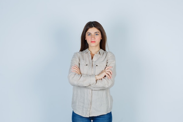 Chica joven en camisa beige, jeans de pie con los brazos cruzados y mirando confiado, vista frontal.