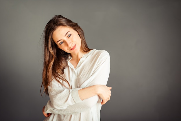 Chica joven en una camisa blanca, manos arriba, en una pared gris. Sin retoques. Sin maquillaje.