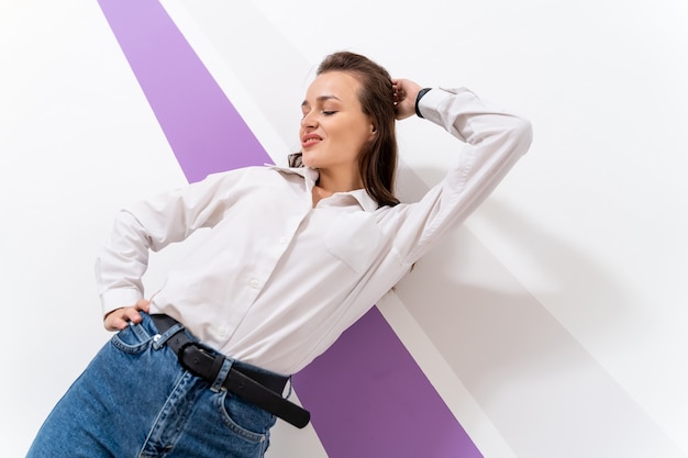 Chica joven con camisa blanca. La mujer está de pie junto a la pared blanca con una mano sobre la cabeza. Blue jeans, estilo casual.