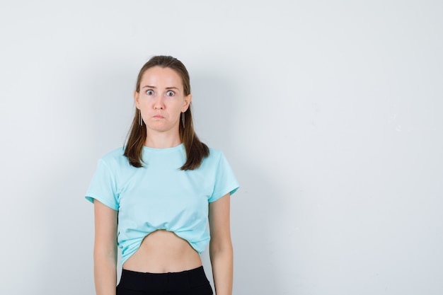 Chica joven en camiseta turquesa, pantalones mirando a cámara, ojos saltones y mirando sorprendido, vista frontal.