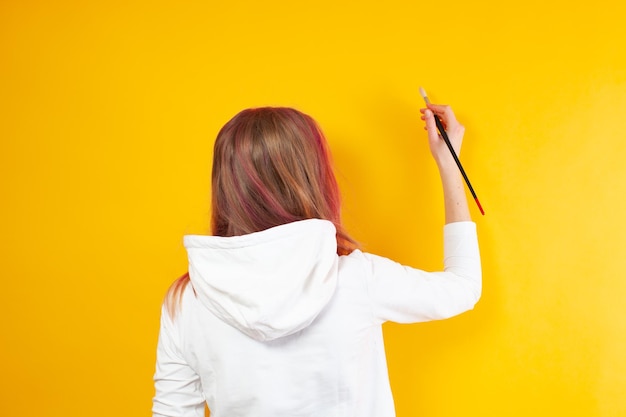 Chica joven con capucha blanca retrovisor escribiendo un mensaje en la pared amarilla usando un lápiz Creando un mapa mental o encontrando una solución al concepto del problema