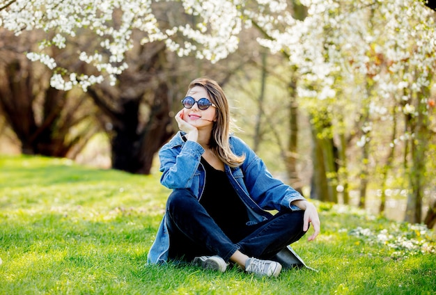 Chica joven con una chaqueta vaquera y gafas de sol sentado cerca de un árbol en flor en el parque. Temporada de primavera