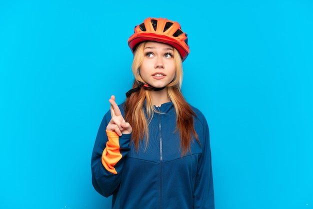 Chica joven ciclista aislada sobre fondo azul con los dedos cruzando y deseando lo mejor