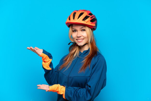 Chica joven ciclista aislada sobre fondo azul extendiendo las manos hacia el lado para invitar a venir