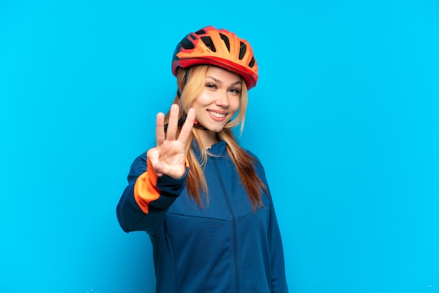 Chica joven ciclista aislada sobre fondo azul feliz y contando tres con los dedos