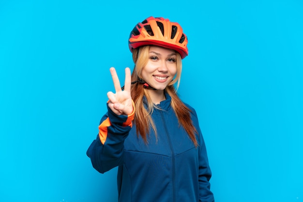 Chica joven ciclista aislada sobre fondo azul sonriendo y mostrando el signo de la victoria