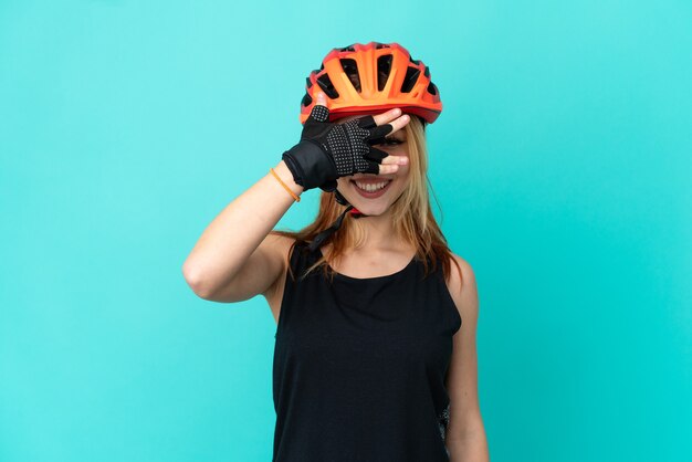 Chica joven ciclista sobre fondo azul aislado cubriendo los ojos con las manos y sonriendo