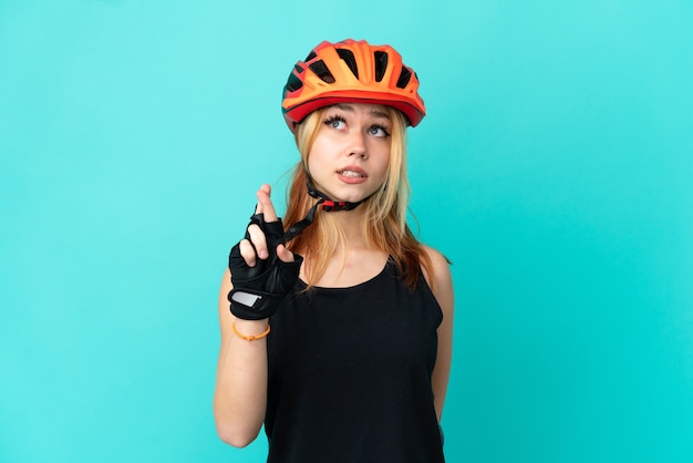 Chica joven ciclista sobre fondo azul aislado con los dedos cruzando y deseando lo mejor