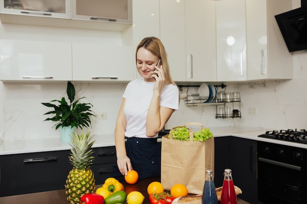 Chica joven en la cocina sosteniendo el teléfono y mirando verduras y frutas en la mesa.