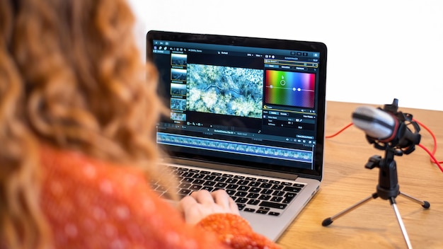 Foto chica joven creadora de contenido editando video en su computadora portátil. micrófono y auriculares sobre la mesa. trabajando desde casa