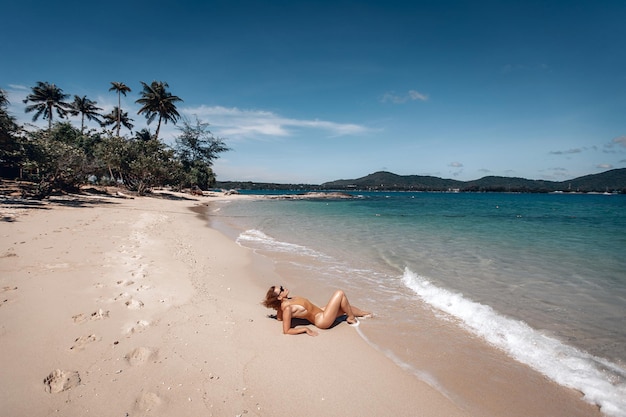 Chica joven con un cuerpo precioso está descansando en la playa con arena blanca cerca del océano. Hermosa modelo sexy en traje de baño beige y gafas de sol negras tomando el sol.