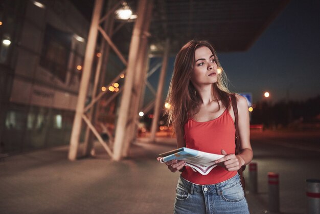 Chica joven cuesta por la noche cerca de la terminal del aeropuerto o estación y lee el mapa de la ciudad