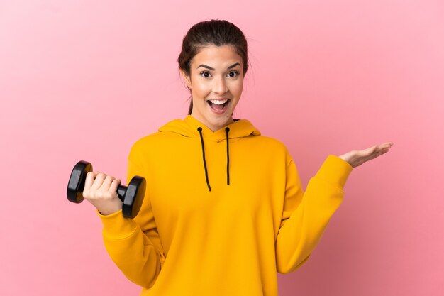 Chica joven deporte haciendo levantamiento de pesas sobre fondo rosa aislado con expresión facial conmocionada