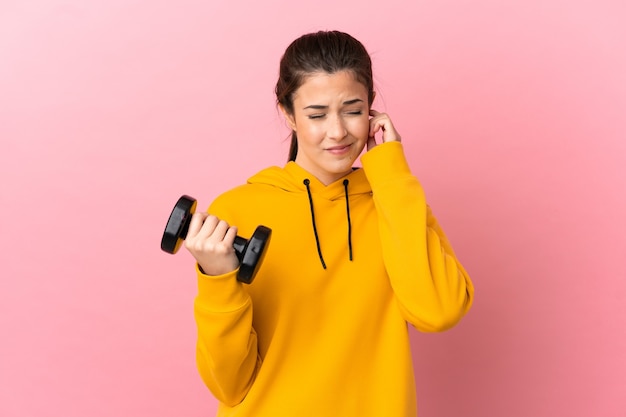 Chica joven deporte haciendo levantamiento de pesas sobre fondo rosa aislado frustrado y cubriendo las orejas