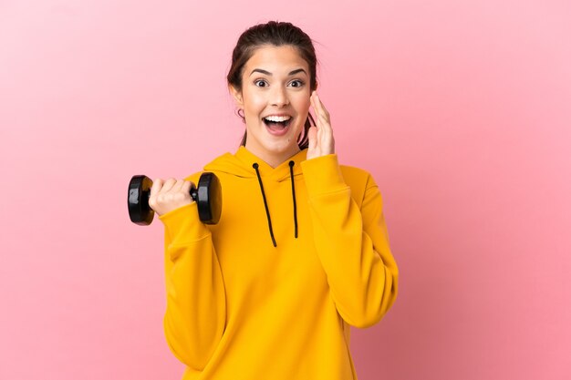 Chica joven deporte haciendo levantamiento de pesas sobre fondo rosa aislado gritando con la boca abierta