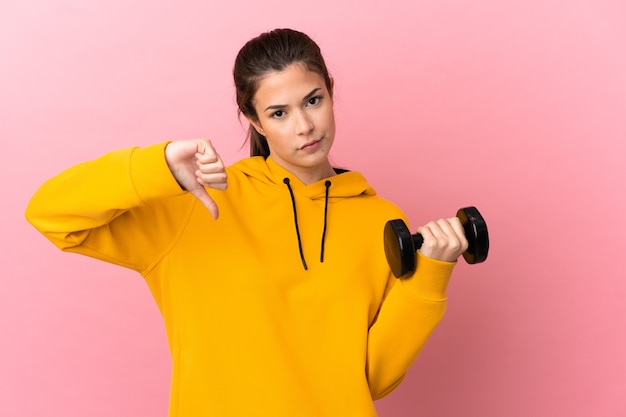 Chica joven deporte haciendo levantamiento de pesas sobre fondo rosa aislado que muestra el pulgar hacia abajo con expresión negativa