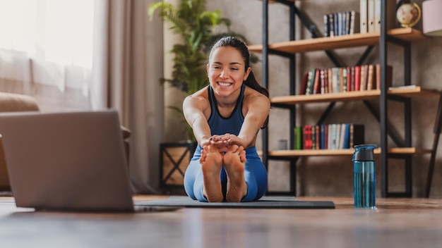 Chica joven deportiva que se estira en la alfombra mientras ve un tutorial en línea en la computadora portátil en casa