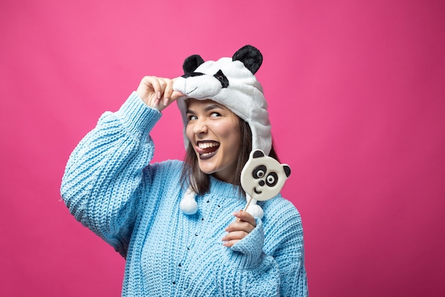Chica joven divertida de pie con un delicioso panda-piruleta en la mano y un sombrero en la cabeza sobre un fondo rosa.