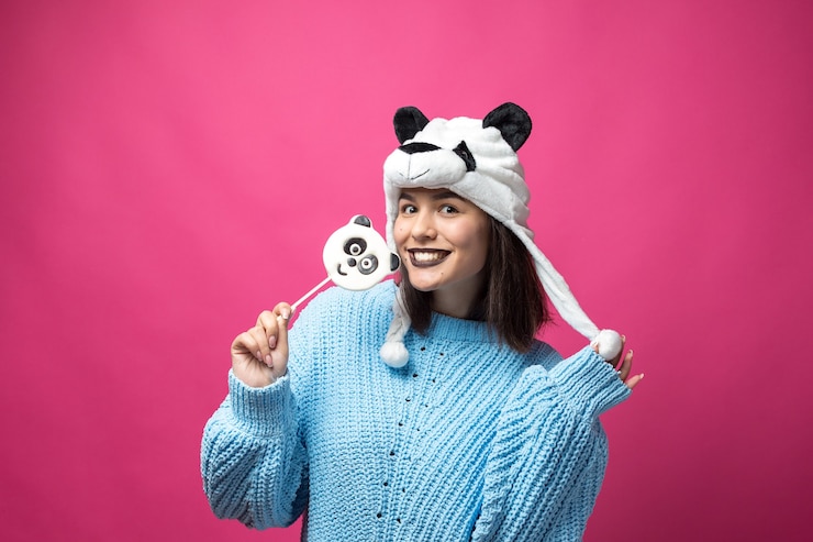 Foto chica joven divertida de pie con delicioso pandalollipop en la mano y un sombrero en la cabeza en un rosa