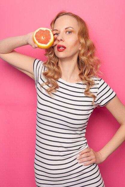 Chica joven divertida en ropa de verano que cubre los ojos con mitades de fruta naranja pomelo madura fresca aislada sobre fondo rosa pastel. La gente viva el estilo de vida relajarse concepto de vacaciones. Simulacros de espacio de copia