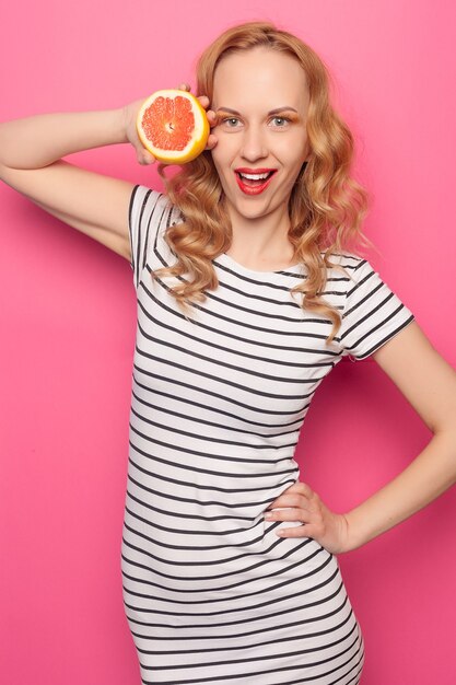 Chica joven divertida en ropa de verano que cubre los ojos con mitades de fruta naranja pomelo madura fresca aislada sobre fondo rosa pastel. La gente viva el estilo de vida relajarse concepto de vacaciones. Simulacros de espacio de copia