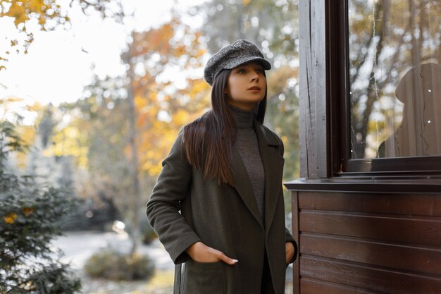 Chica joven y elegante con un abrigo gris y una gorra con estilo se encuentra en el parque en una tarde de otoño. Mujer hermosa