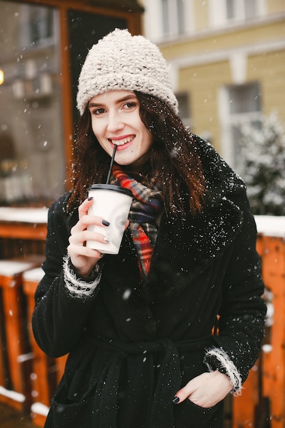 chica joven y elegante en abrigo negro y sombrero blanco tomando café en la ciudad de invierno