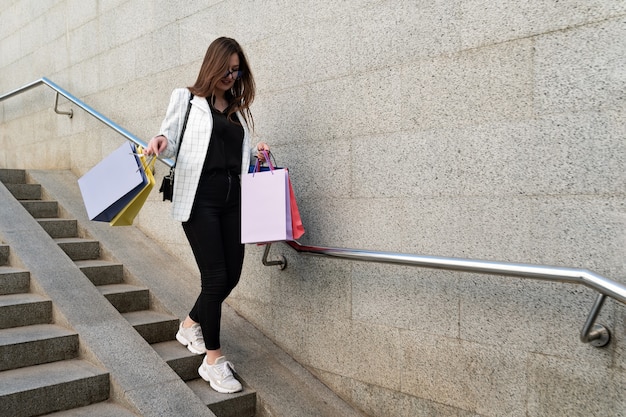 Chica joven elegante con bolsas multicolores después de ir de compras baja las escaleras.