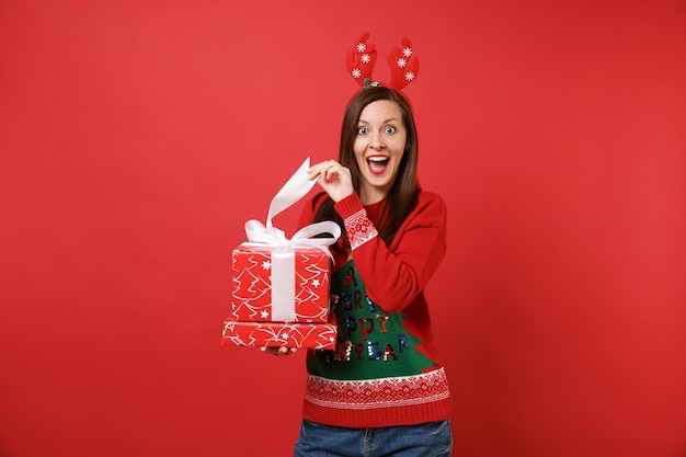 Chica joven emocionada de Santa en divertidos cuernos de ciervo decorativos en la cabeza abriendo cajas rojas con regalo presente aislado sobre fondo rojo. Feliz año nuevo 2019 celebración concepto de fiesta navideña. Simulacros de espacio de copia.