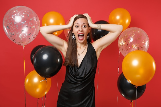 Chica joven emocionada en vestido negro gritando, celebrando, poniendo las manos en la cabeza en globos de aire de fondo rojo brillante. Día internacional de la mujer, concepto de fiesta de vacaciones de maqueta de cumpleaños de feliz año nuevo.