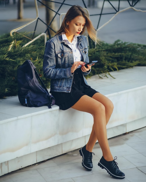 Chica joven en entorno urbano utiliza smartphone Ropa casual chaqueta de mezclilla falda corta Imagen vertical