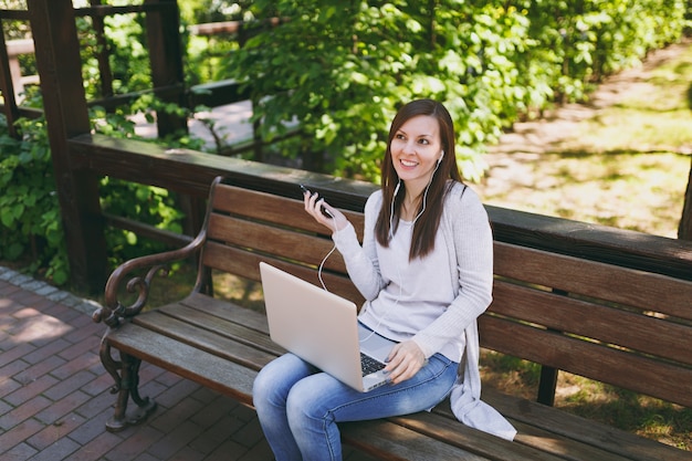 Chica joven escucha música en el teléfono móvil con auriculares. Mujer sentada en un banco trabajando en un moderno ordenador portátil en el parque de la ciudad en la calle al aire libre en la naturaleza. Oficina móvil. Concepto de negocio autónomo