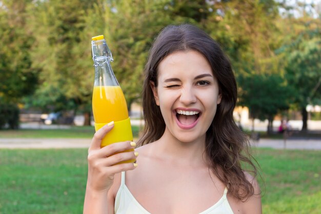 La chica joven está caminando en el parque que sostiene una botella de jugo fresco o de limonada en un día cálido y soleado