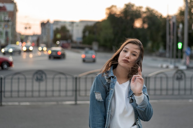 La chica joven está posando junto a la carretera. Retrato de estudiante en el fondo de la ciudad. Mujer joven con estilo.