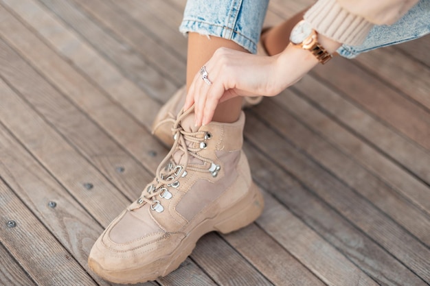 Chica joven con estilo en botas de invierno beige de moda ata los cordones de los zapatos sobre un fondo de madera, primer plano