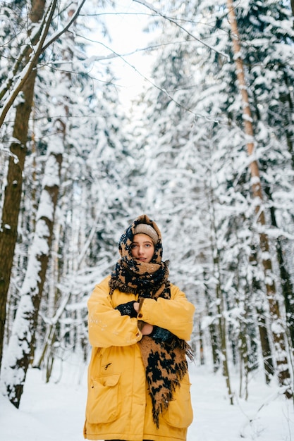 Chica joven con estilo hipster en chaqueta amarilla que cubre la cabeza con un pañuelo caliente y posando en el bosque