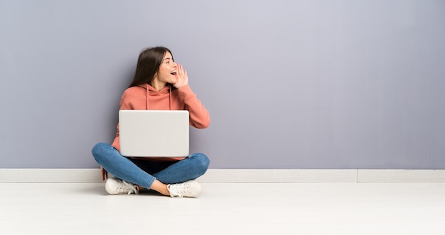 Chica joven estudiante con una computadora portátil en el piso gritando con la boca abierta