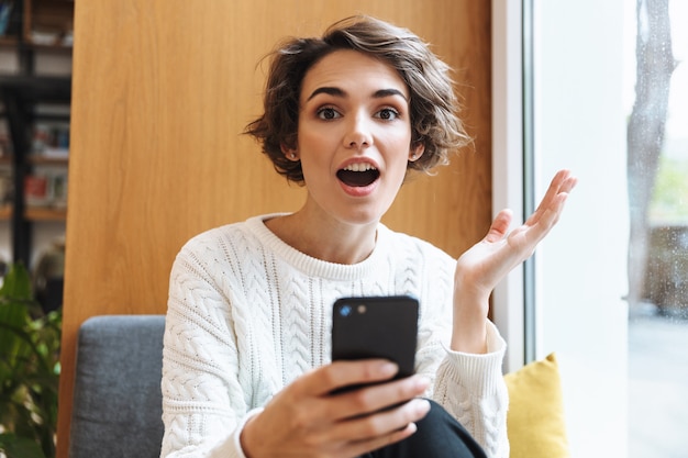 Chica joven estudiante feliz estudiando en la biblioteca, mediante teléfono móvil