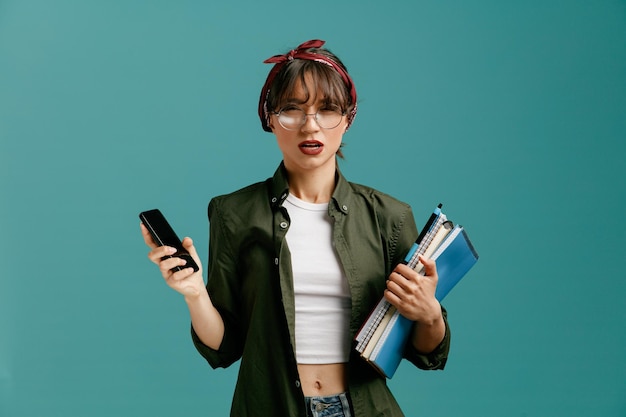 Chica joven estudiante insegura con gafas de bandana sosteniendo un teléfono móvil y grandes blocs de notas mirando a la cámara aislada en el fondo azul