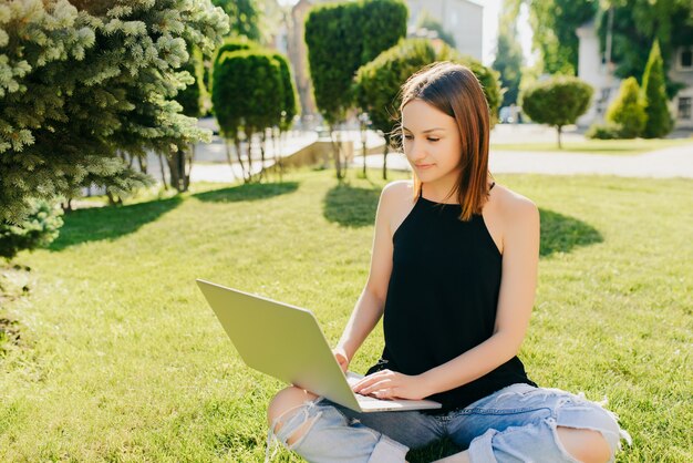 Chica joven estudiante en ropa casual trabajando en una computadora portátil, estudiando, preparándose para los exámenes, mientras está sentado en el césped, en el parque.